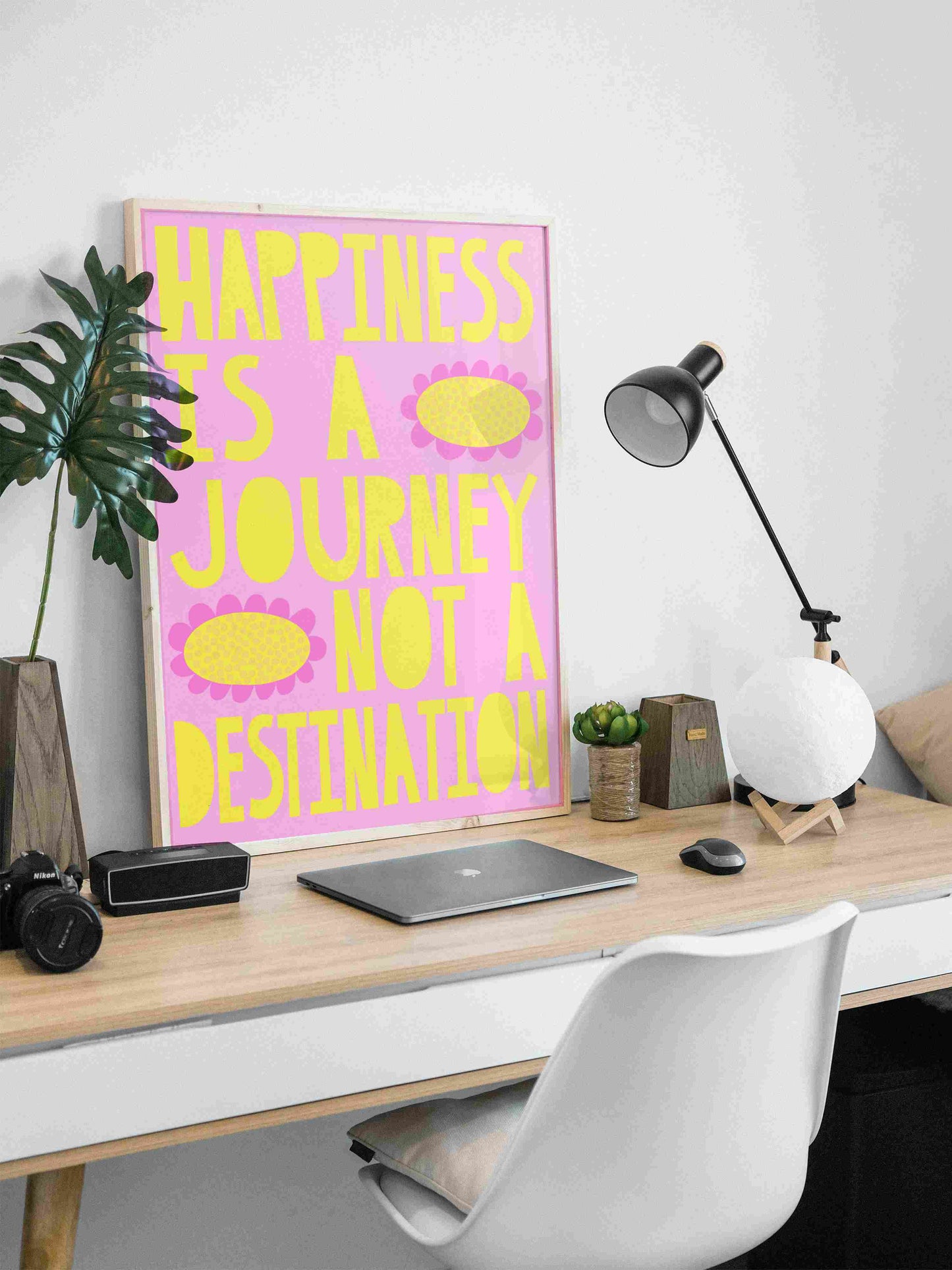 This is an image of what this poster would look when printed. It is in a frame that is sitting on a desk. The artwork has a bright lolly pink background with bold yellow words that read "Happiness is a journey not a destination." There are two bold flowers that have dark pink petals and yellow centres.